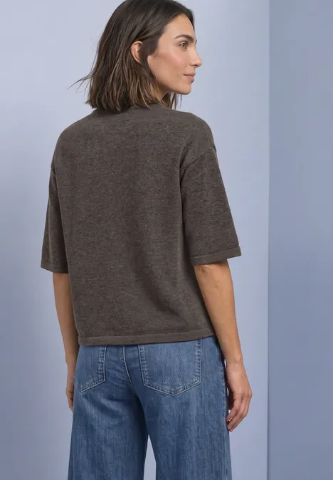 Frau von hinten in braunem Kurzarm-Pullover und blauen Jeans vor hellgrauem Hintergrund.