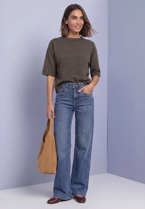 Eine Frau mit kurzem Haar trägt ein khakifarbenes Shirt, weite Jeans, braune Schuhe und hält eine Tasche.