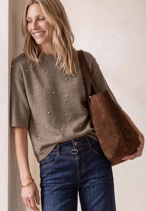 Eine lächelnde Frau trägt einen Perlenpullover, Jeans und eine braune Wildledertasche.