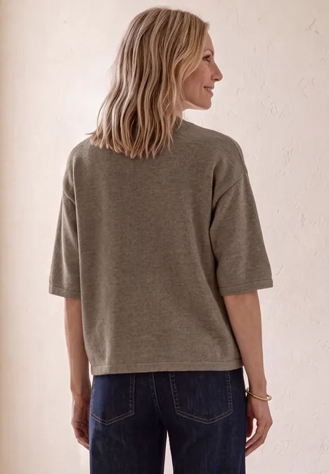 Frau von hinten in grauem, lockerem Oberteil und blauen Jeans vor hellem, neutralem Hintergrund.