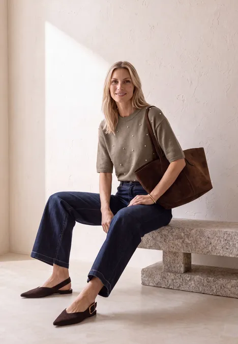 Blonde Frau in grünem Pullover, Jeans und brauner Tasche sitzt auf einer Bank in hellem Raum.
