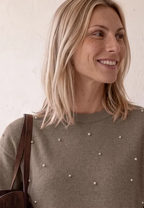 Eine blonde Frau mit Perlen-Verzierung am Pullover trägt eine Tasche und lächelt sanft seitlich.