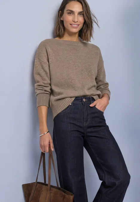 Frau in kastanienbraunem Pullover und dunkler Jeans, hält eine braune Handtasche, steht vor hellem Hintergrund.