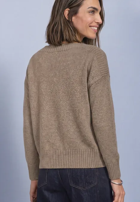 Frau in braunem Pullover und Jeans, Rückansicht, leicht zur Seite schauend, lächelnd vor hellem Hintergrund.