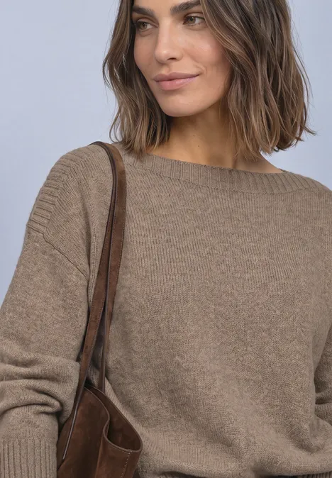 Frau mit schulterlangen Haaren trägt braunen Pullover und passende Tasche, lächelt seitlich vor neutralem Hintergrund.