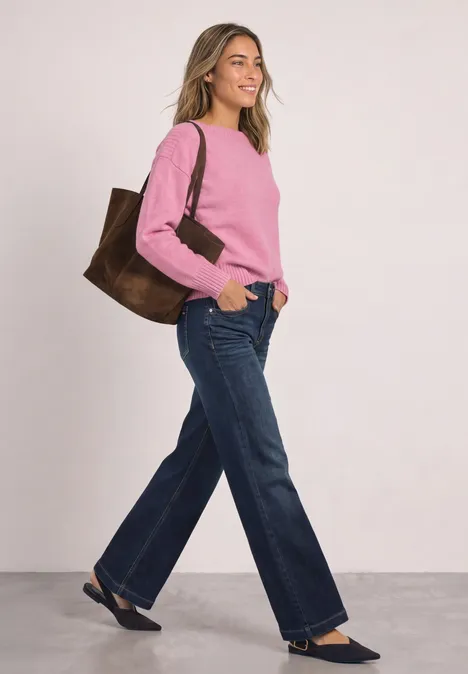 Frau mit rosa Pullover, weiter Jeans, schwarzen Flats und großer brauner Tasche posiert lächelnd.