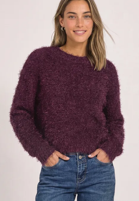 Jonge vrouw draagt een donzige paarse trui en een blauwe jeans voor een lichte achtergrond.