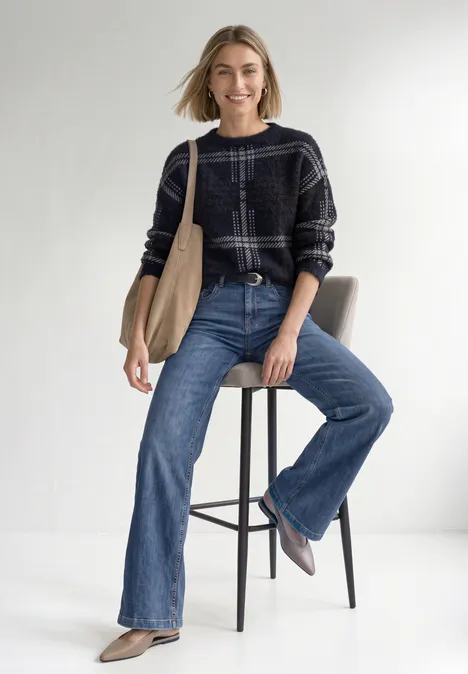 Langarm Pullover mit Karomuster indigo night Frau sitzt auf Hocker, lächelt, trägt weite Jeans, kariertes Hemd und Schultertasche.