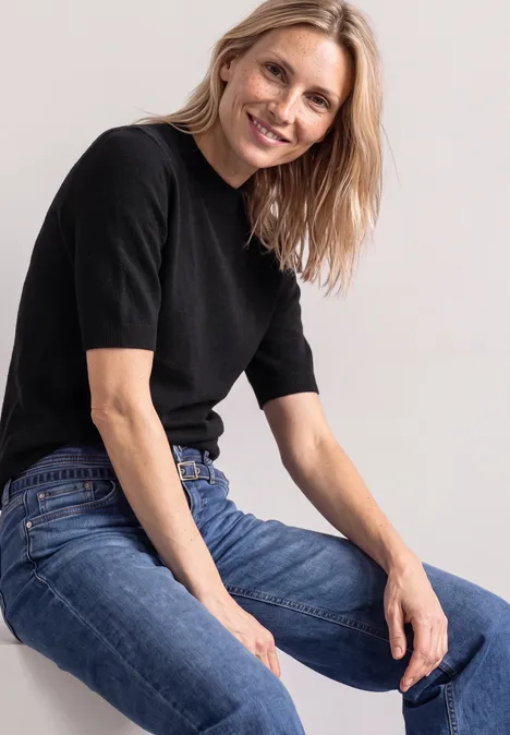 Eine blonde Frau in schwarzem T-Shirt und Jeans sitzt lächelnd vor hellem Hintergrund.