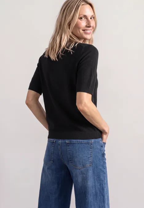 Eine Frau steht mit dem Rücken zur Kamera, schaut über die Schulter, trägt ein schwarzes Shirt und blaue Jeans.