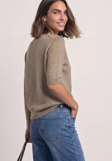 Frau in modischem, hellbraunem Kurzarm-Pullover und blauen Jeans, schaut über die Schulter und lächelt.