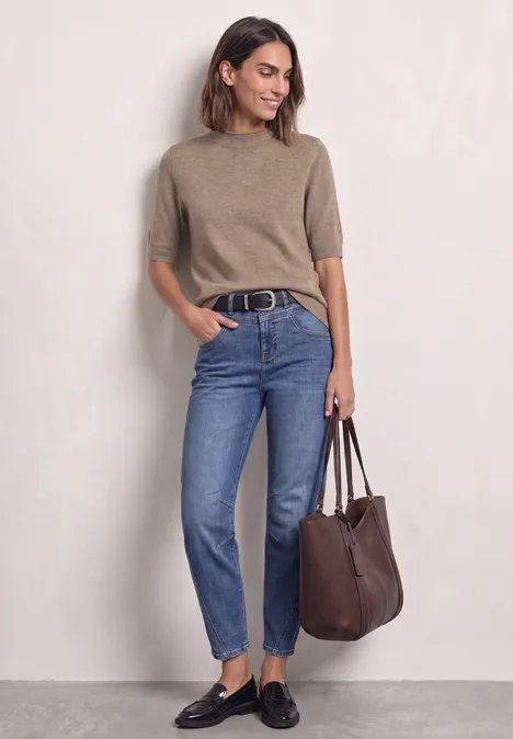 Frau in beigem Pullover, blauen Jeans und schwarzen Loafers hält eine braune Tasche.