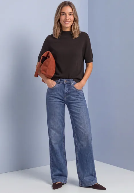 Vrouw in zwart T-shirt en wijde jeans draagt een bruine trui over haar schouder voor een lichtblauwe achtergrond.