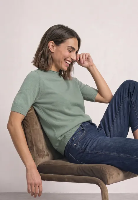 Vrouw in groene trui en spijkerbroek, zittend op een stoel, lacht.