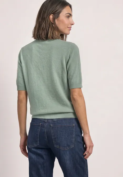 Vrouw in groen kort mouwtruitje en jeans, van opzij met de rug naar de camera tegen neutrale achtergrond.