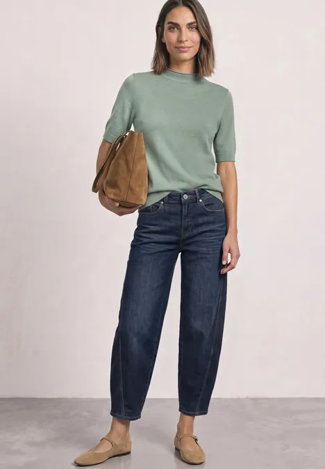 Vrouw met olijfgroene trui, donkerblauwe jeans, beige schoenen en bruine tas tegen neutrale achtergrond.