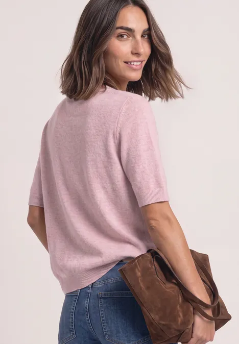 Vrouw in roze korte-mouwtrui en blauwe jeans, met een bruine tas in haar hand, licht zijwaarts pose.
