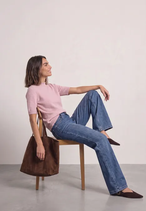 Vrouw zit zijwaarts op een stoel, roze top, blauwe jeans, bruine schoenen, bruine jas over de leuning.