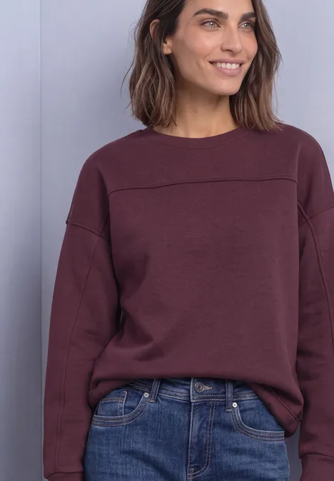 Eine Frau in einem weinroten Pullover und Jeans, lächelt vor neutralem grauem Hintergrund.