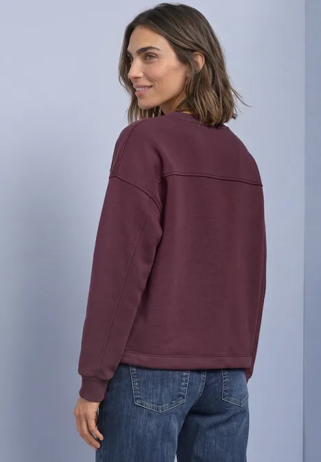 Frau in bordeauxrotem Sweatshirt und blauen Jeans, schaut über die Schulter vor hellem Hintergrund.