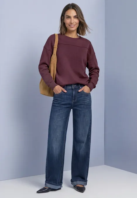 Frau in lila Pullover, weiten Jeans und Handtasche lächelnd vor neutralem Hintergrund.