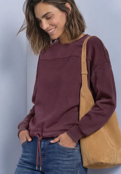 Lächelnde Frau in lila Sweatshirt, Jeans und hellbrauner Schultertasche vor hellem Hintergrund.