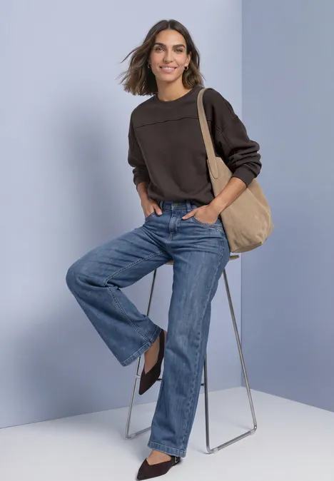 Frau in braunem Pullover und weitem Jeans, mit Tasche, lehnt locker an einem Hocker vor schlichtem Hintergrund.