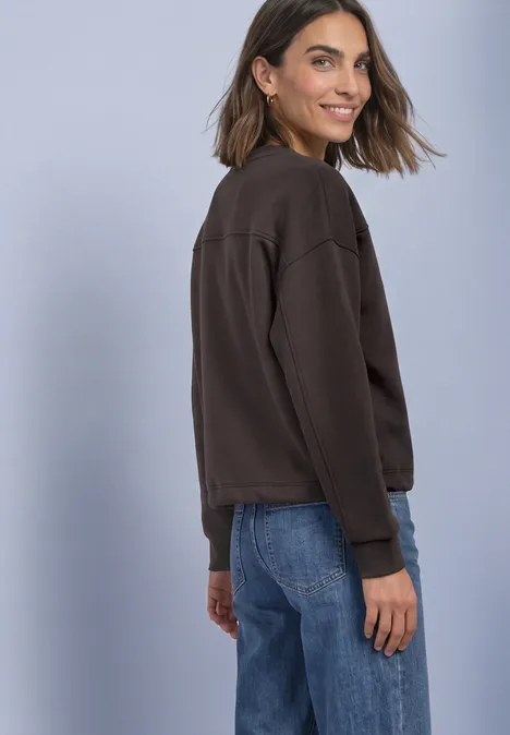 Eine lächelnde Frau in braunem Pullover und Jeans, schaut über die Schulter vor hellem Hintergrund.