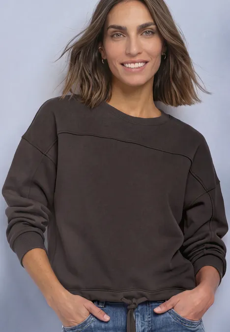 Frau mit lässigem Pullover in dunkler Farbe, leicht lächelnd vor neutralem Hintergrund.