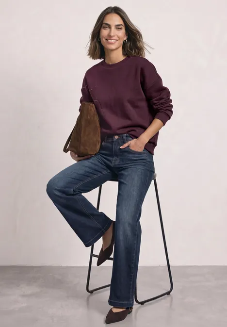 Frau in Jeans und weinrotem Pullover sitzt lässig auf einem Hocker, hält eine braune Tasche.