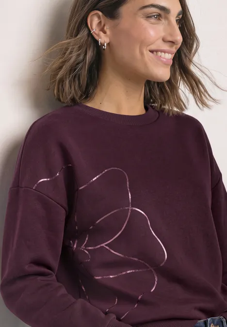 Frau lächelt seitlich in einem burgunderfarbenen Pullover mit dezentem Blumenlinienmuster.