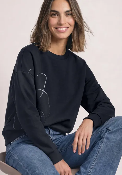 Frau mit schwarzem Sweatshirt mit Linienzeichnung und blauen Jeans, lächelt in die Kamera.