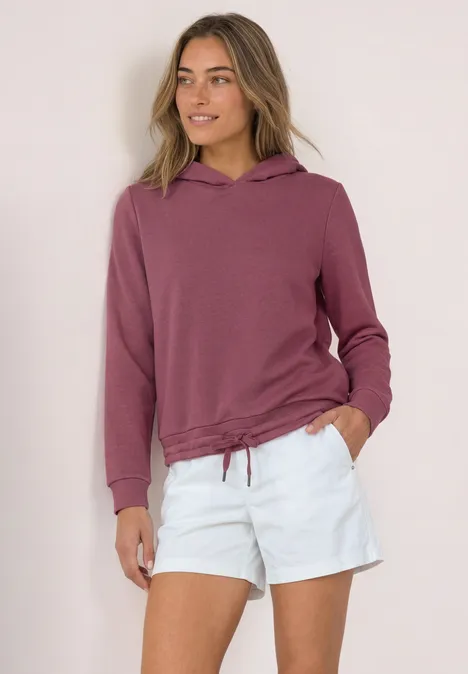Frau in mauvefarbenem Hoodie mit Tunnelzug und weißen Shorts, steht entspannt mit Händen in den Taschen.