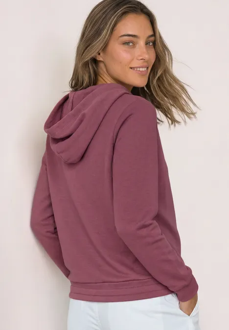Eine Frau trägt einen weinroten Kapuzenpullover und schaut lächelnd über die Schulter.