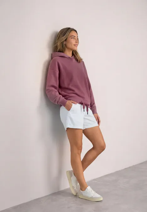 Frau lehnt lässig an der Wand in rosa Hoodie, weißen Shorts und Sneakers.
