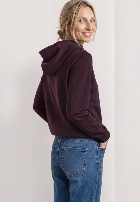 Lächelnde Frau in violetter Kapuzenjacke und Jeans, seitlich mit Händen in den Taschen.