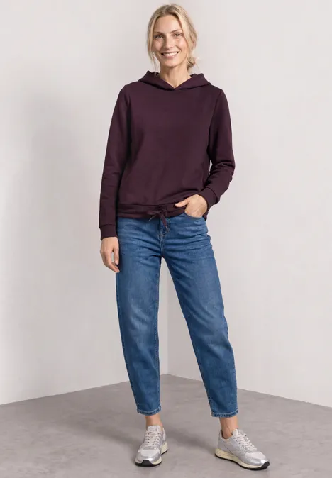 Lächelnde Frau in lila Kapuzenpullover, blauen Jeans und Sneakers vor neutralem Hintergrund.