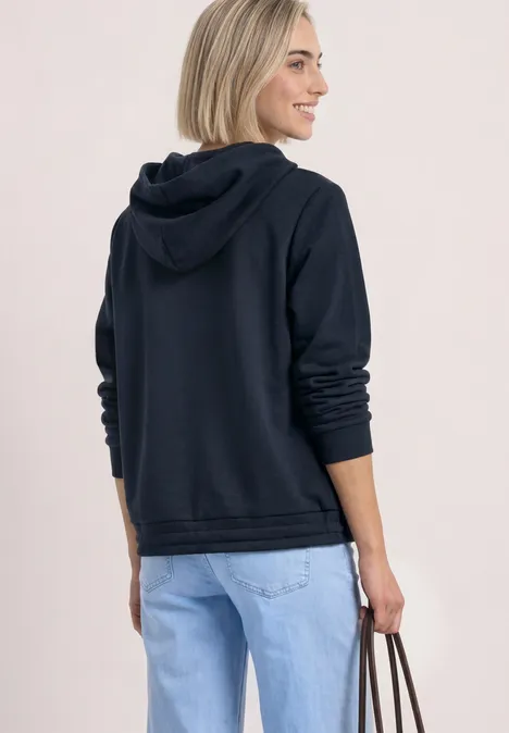 Eine Frau mit kurzem hellblonden Haar trägt eine schwarze Kapuzenjacke, helle Jeans und hält eine Tasche.