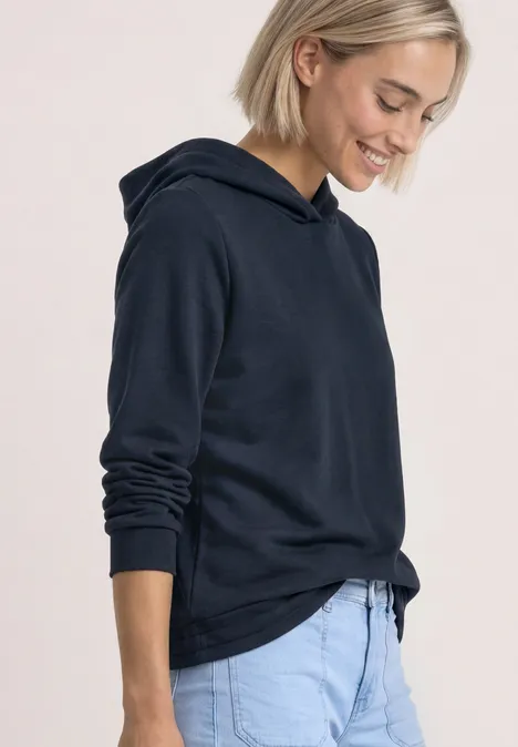 Eine Frau in dunkelblauem Hoodie und hellblauen Hosen lächelt seitlich.