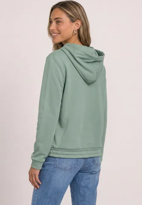 Junge Frau trägt mintgrünen Hoodie und Jeans, blickt lächelnd über die Schulter vor hellem Hintergrund.