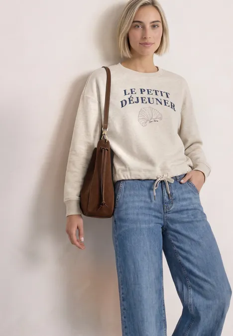 Frau trägt beigen Pullover mit 'Le Petit Déjeuner'-Schriftzug, weite Jeans und braune Tasche vor hellem Hintergrund.