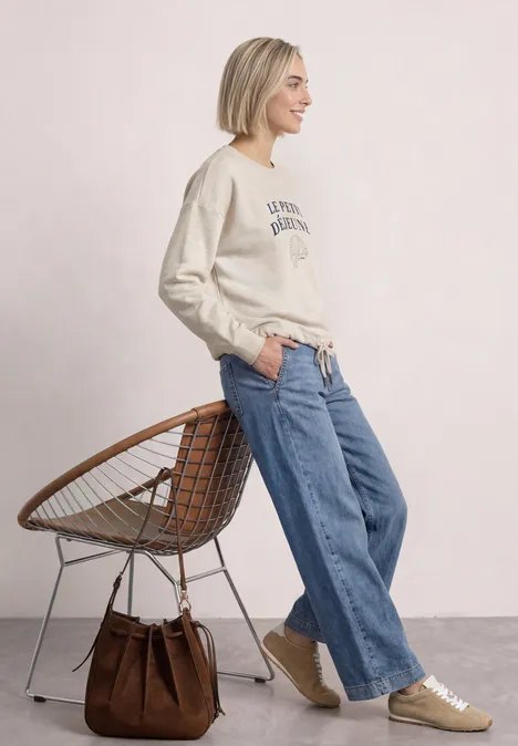Frau in beigem Sweatshirt, weiten Jeans und Sneakern steht lässig neben einem Korbstuhl, braune Tasche.