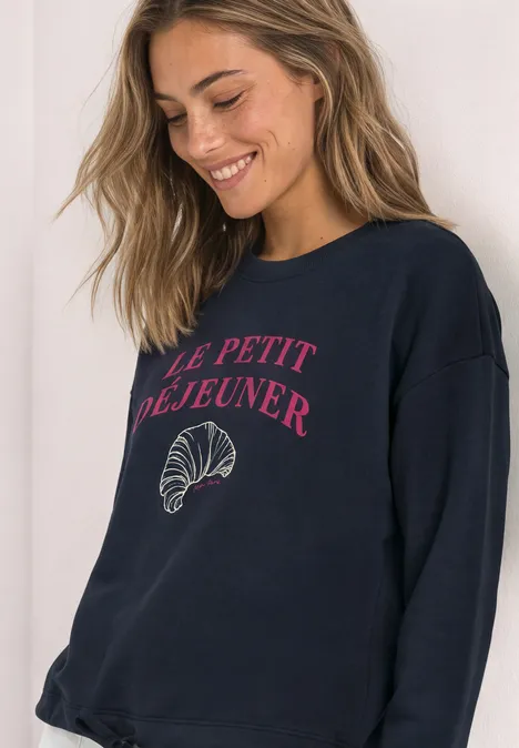 Frau in dunkelblauem Sweatshirt mit rosafarbener Aufschrift «Le petit déjeuner» und Croissant-Motiv lächelt.