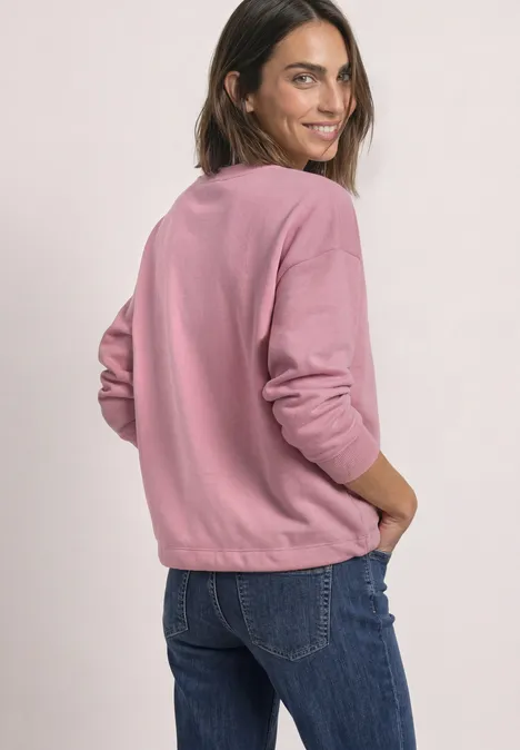 Eine Frau in rosa Sweatshirt und blauer Jeans blickt lächelnd über die Schulter.