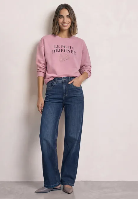 Frau in rosa Pullover mit Aufschrift 'Le petit déjeuner' und weiten blauen Jeans