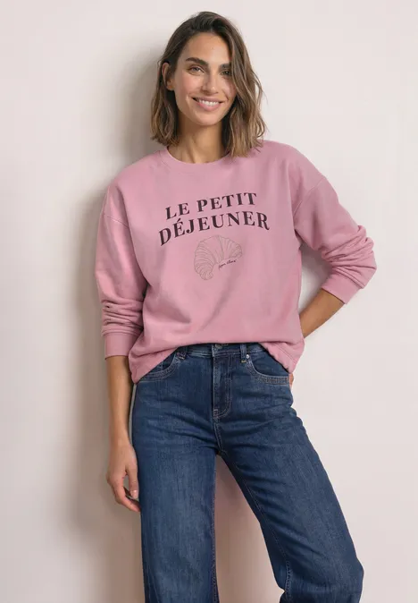 Frau in Jeans und rosa Sweatshirt mit Aufschrift „Le Petit Déjeuner“, lächelnd mit einer Hand in der Tasche.