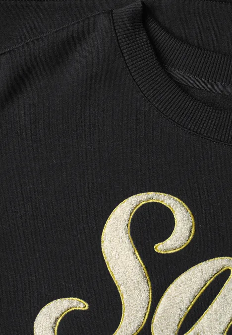 Schwarzes Oberteil mit funkelndem silbernem Glitzerbuchstaben „S“. 