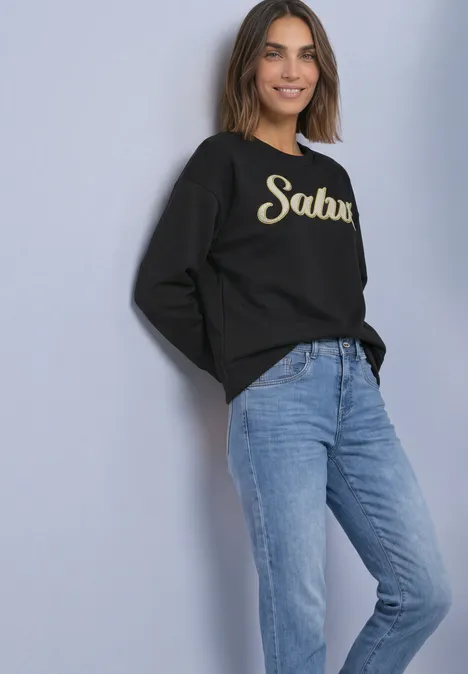 Frau lehnt an Wand, trägt schwarzen Pullover mit 'Salut'-Aufdruck und blaue Jeans, lächelnd.
