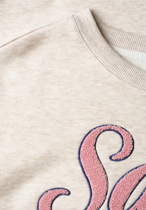 Nahaufnahme eines beigen Sweatshirts mit rosa, flauschigem Buchstaben-Patch.