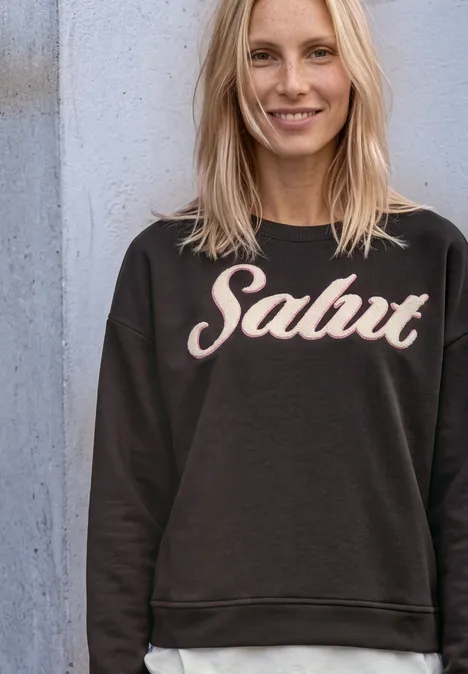Sweatshirt met lange mouwen en Artwork black coffee Fröhliche blonde Frau in schwarzem Pullover mit der Aufschrift 'Salut', lehnt lächelnd an einer Wand.